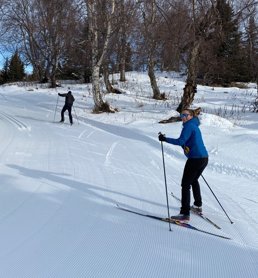 IMG_3669 a small girl with a mask on skis and in winter clothing, standing in front of her moth on skis on a snowy trail